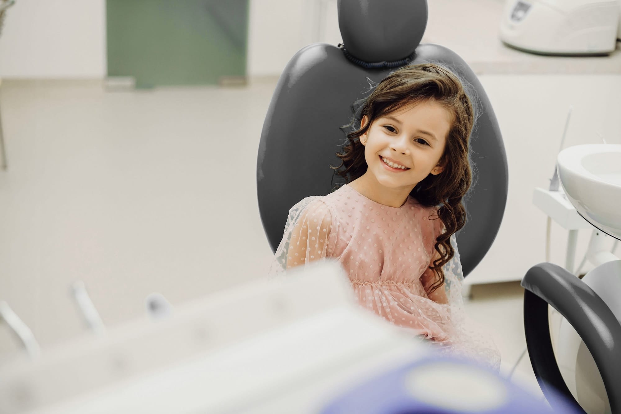 Kind im Zahnarztstuhl: Ein Mädchen sitzt entspannt im Behandlungsraum und schaut freundlich in die Kamera.