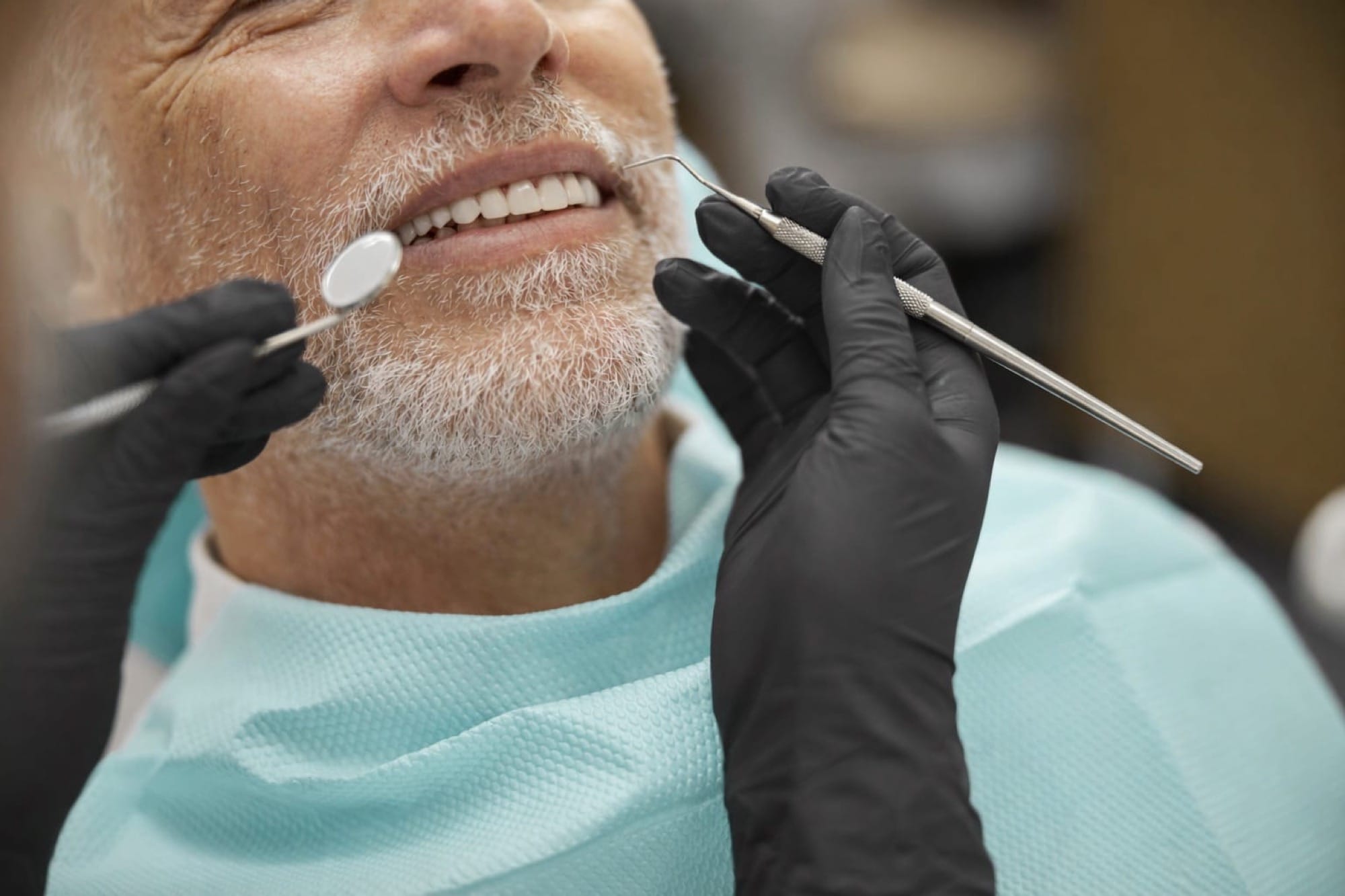 Zahnuntersuchung bei Mann: Ein älterer Mann sitzt im Stuhl, während der Zahnarzt mit Spiegel und Instrumenten die Zähne kontrolliert.